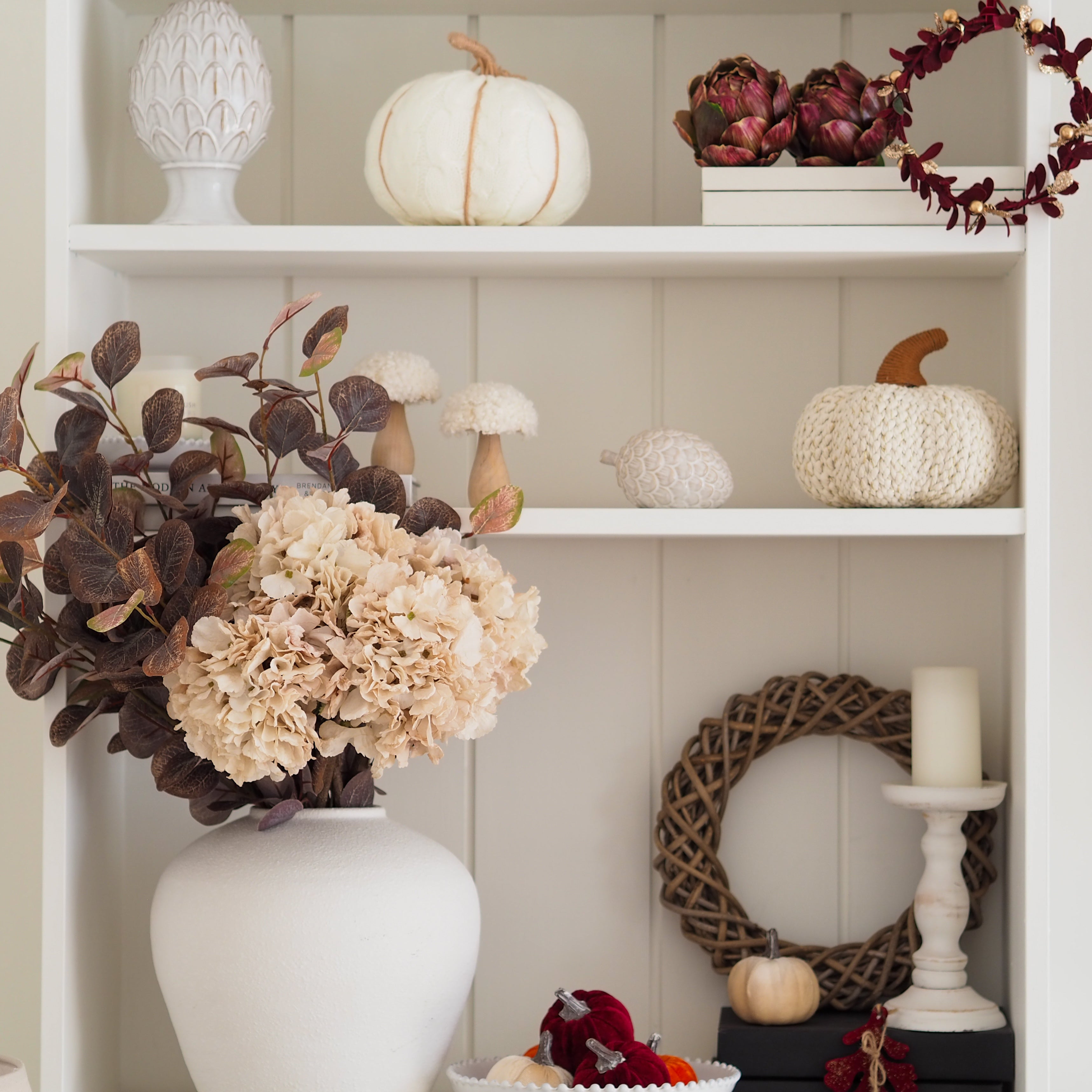 A styled autumn shelf display featuring cream and white pumpkins, a white ceramic vase filled with beige hydrangea blooms and dark brown leaves, a woven wreath, a candle on a white holder, and decorative artichokes against a white paneled background. Home Decor