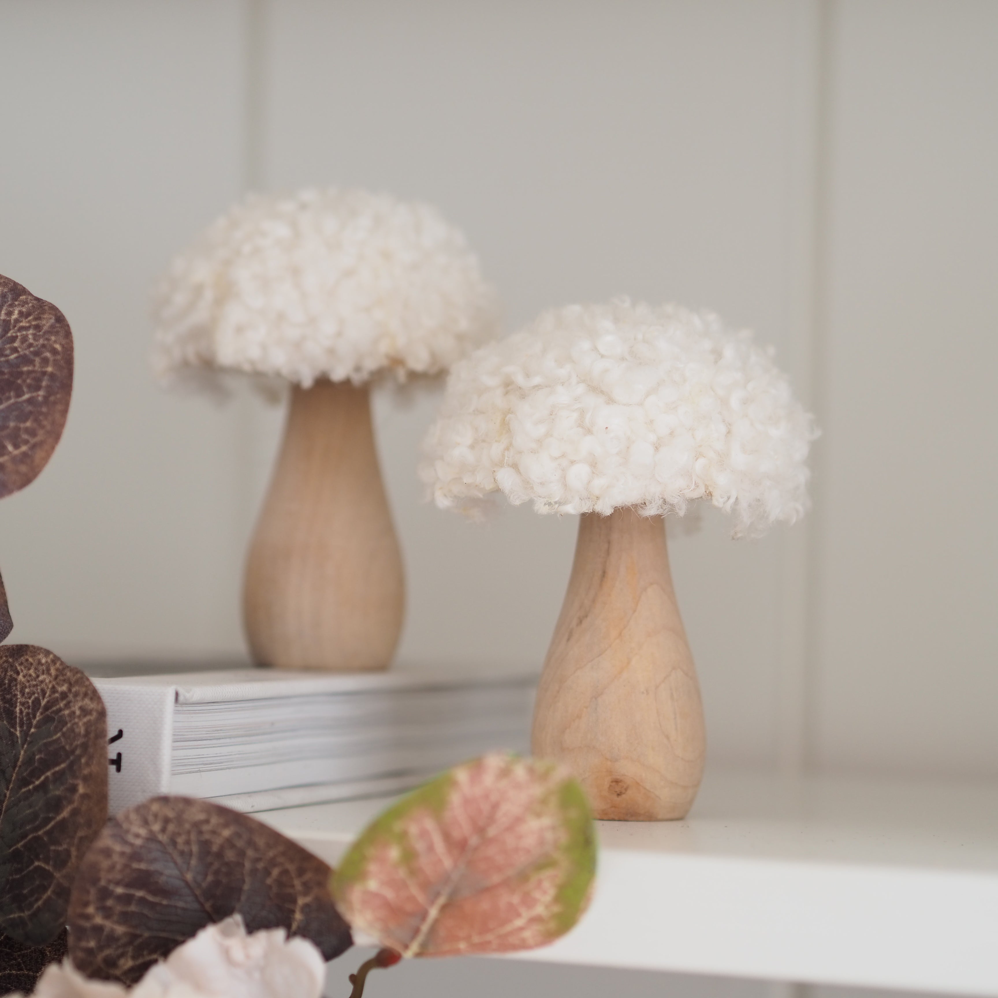 Two decorative mushroom-shaped objects with fluffy off-white caps and light natural wood bases, displayed on a white shelf with dried leaves in the foreground.