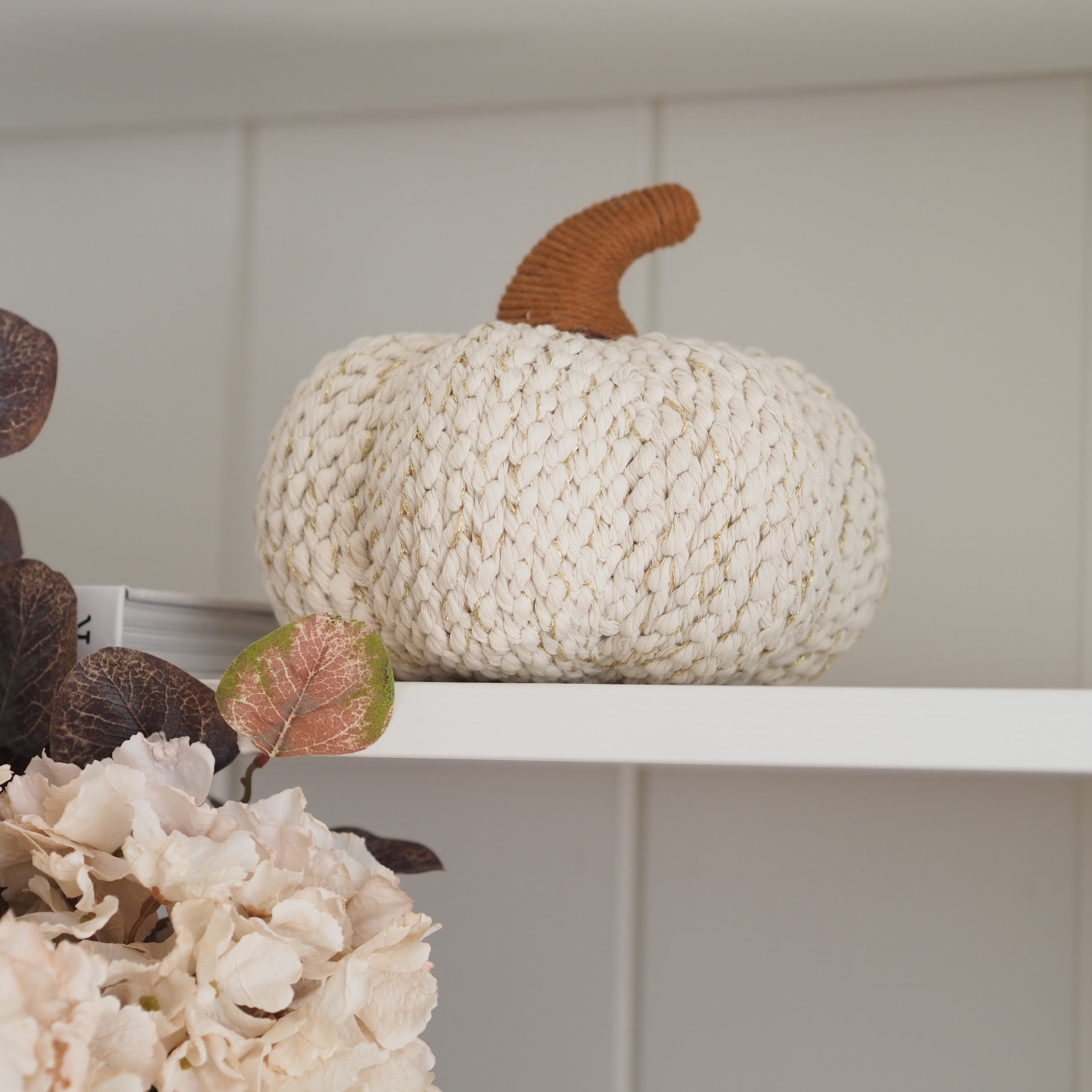 A chunky knit decorative pumpkin in cream with a brown stem, displayed on a white shelf next to dried leaves and pale flowers.