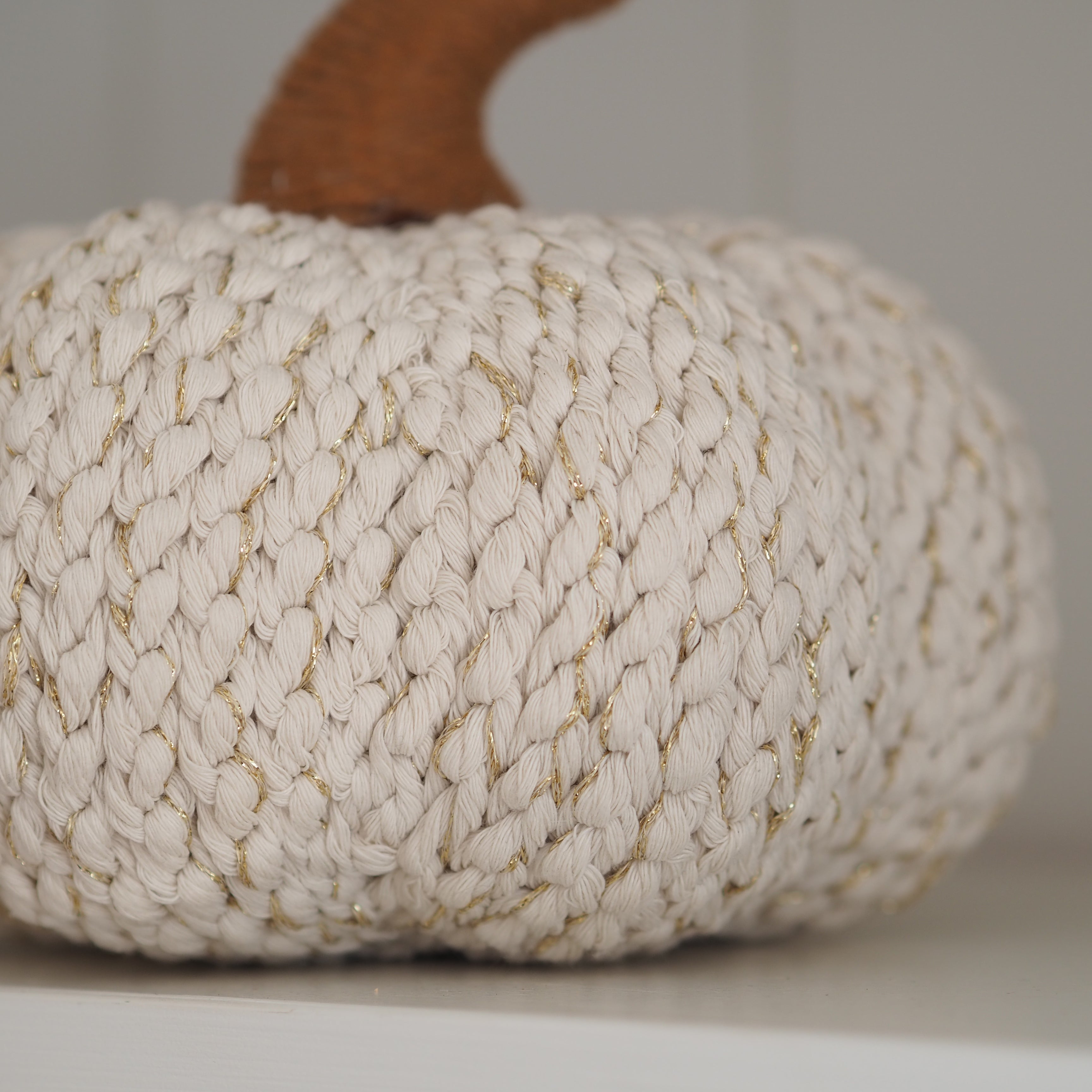 A close-up of a decorative rope pumpkin with a chunky off-white woven body, subtle gold thread accents, and a brown stem, resting on a light surface against a neutral background.