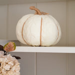 A cream-colored knit pumpkin decoration with a natural jute stem, displayed on a white shelf next to dried leaves and pale flowers.