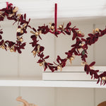 A heart-shaped wreath made of deep burgundy velvet leaves and gold glitter accents, hanging from a white shelf.