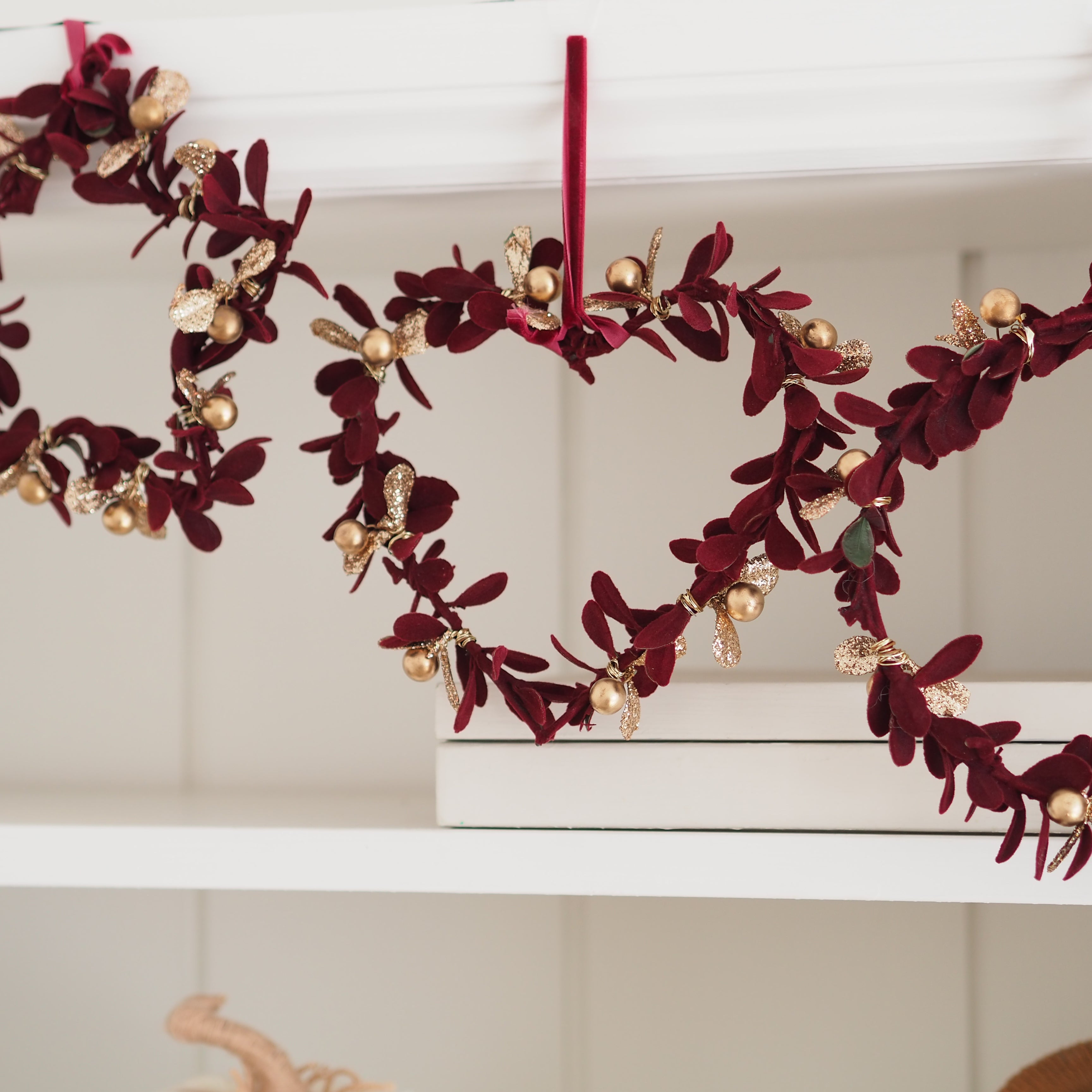 A heart-shaped wreath made of deep burgundy velvet leaves and gold glitter accents, hanging from a white shelf.