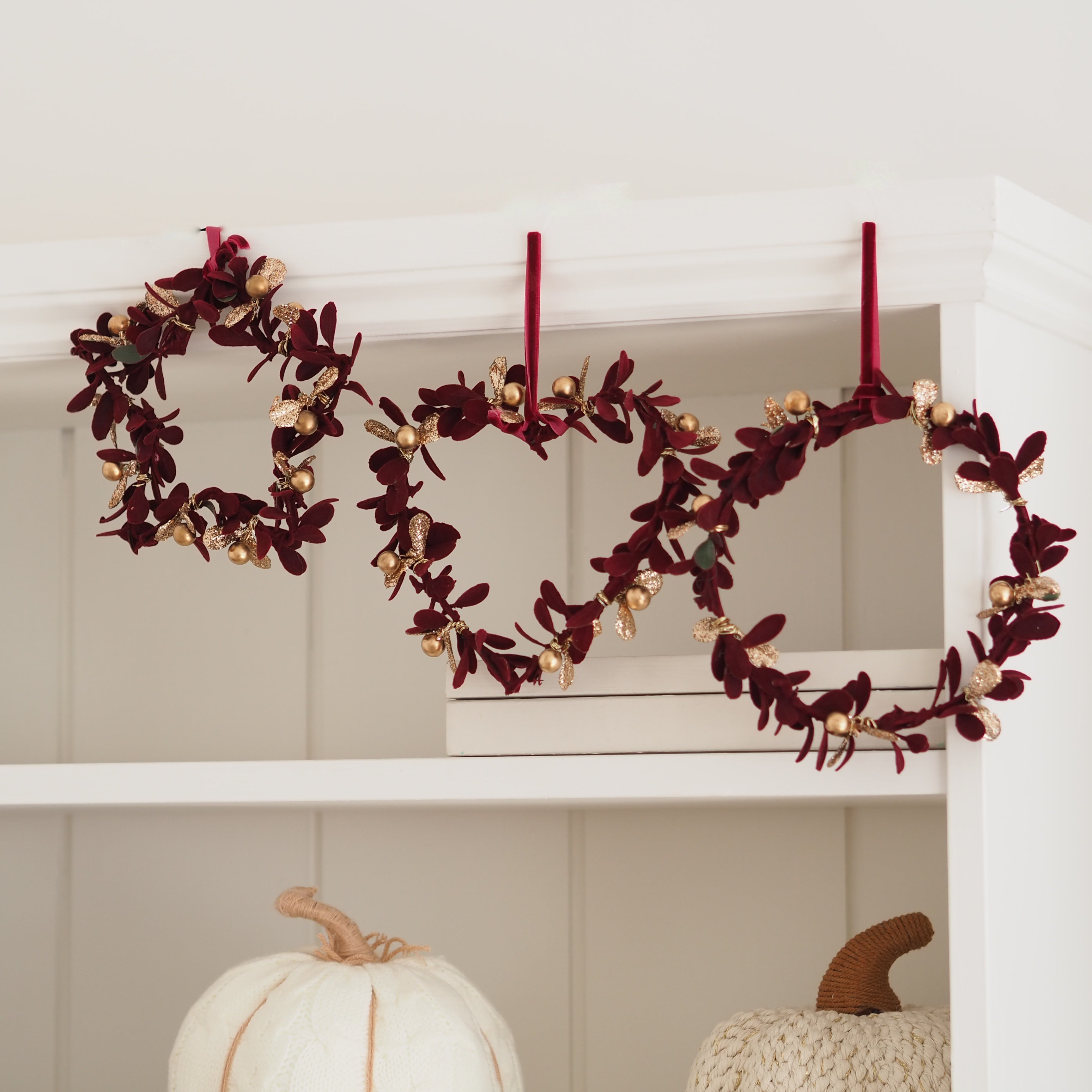 Three autumn-themed wreaths with burgundy leaves and gold pinecones hang from red ribbons on a white shelf above stacked white books, with decorative pumpkins below.
