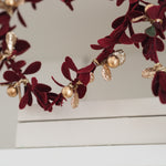 A close-up of a decorative wreath with deep burgundy velvet leaves, gold metallic accents, and small pearl-like gold beads, draped over a white surface against a light neutral background.