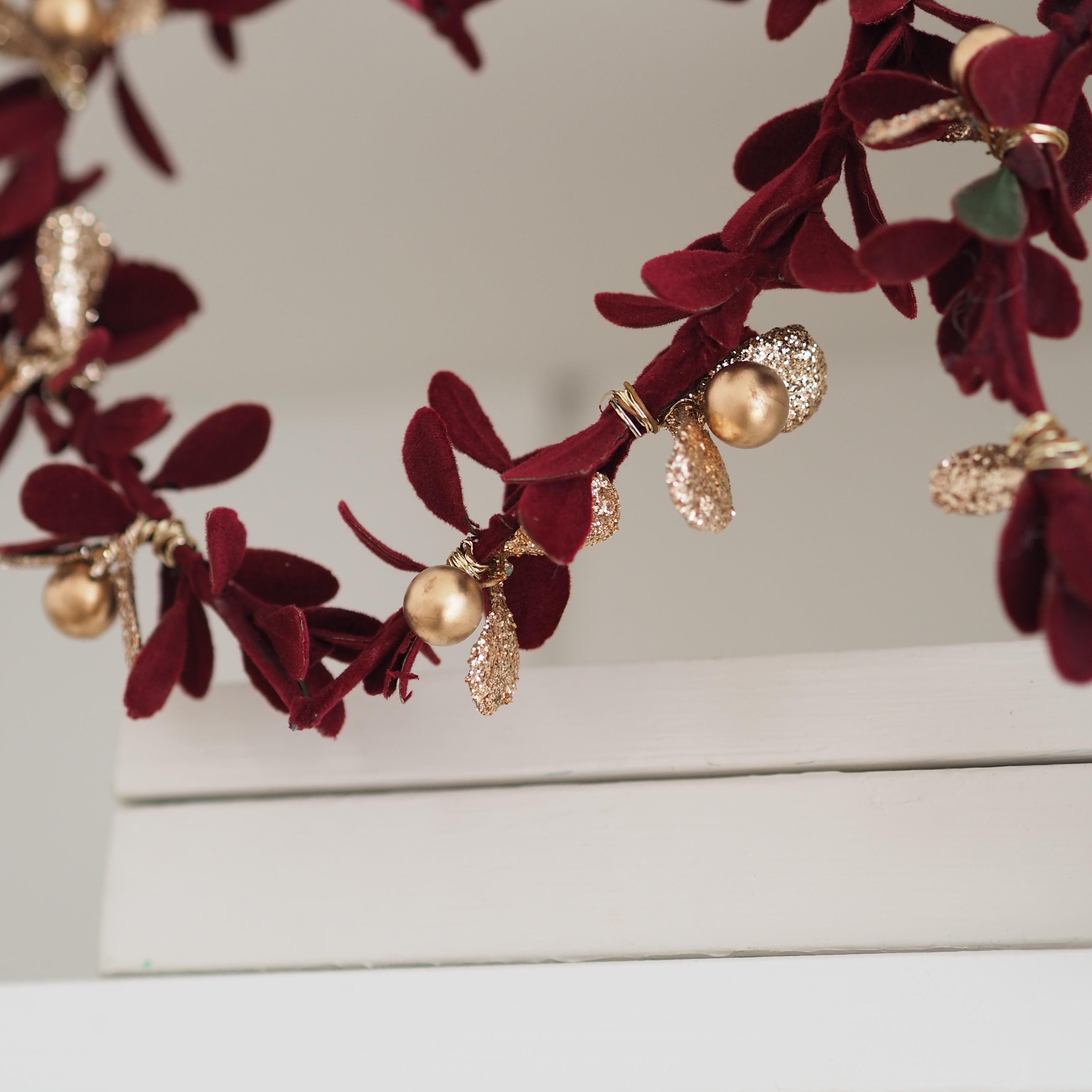 A close-up of a decorative wreath with deep burgundy velvet leaves, gold metallic accents, and small pearl-like gold beads, draped over a white surface against a light neutral background.