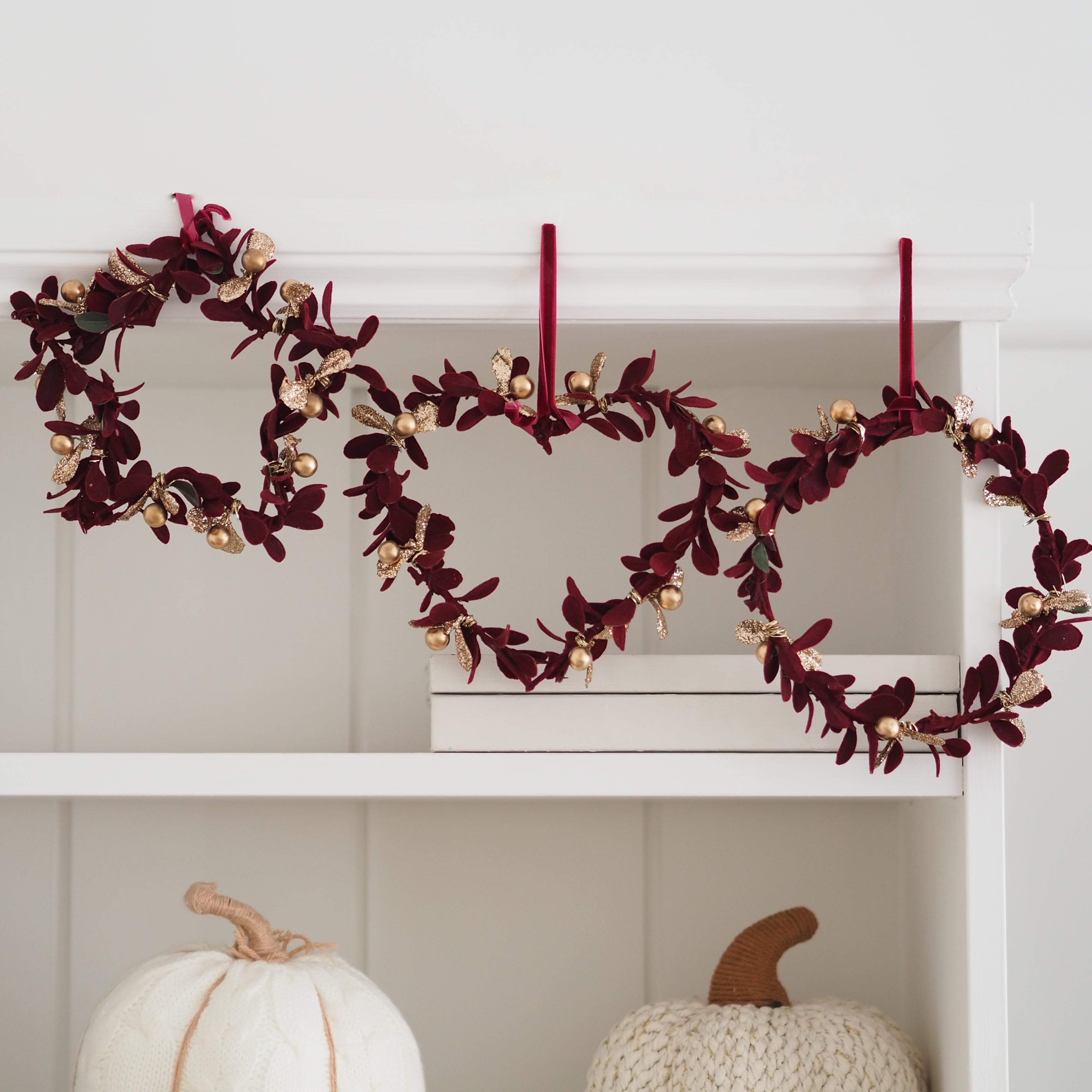 A fall-themed garland with burgundy leaves and gold berries hangs across a white shelf, flanked by white and cream pumpkins below.