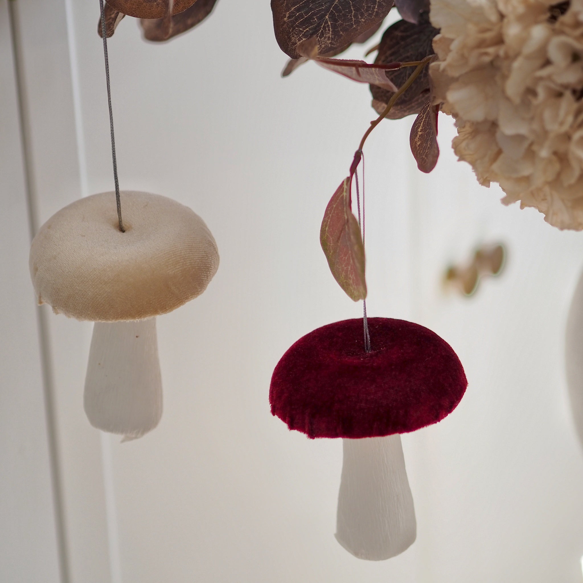 Two mushroom-shaped hanging ornaments, one in cream and one in deep burgundy velvet, suspended against a soft white background with dried leaves and cream flowers.