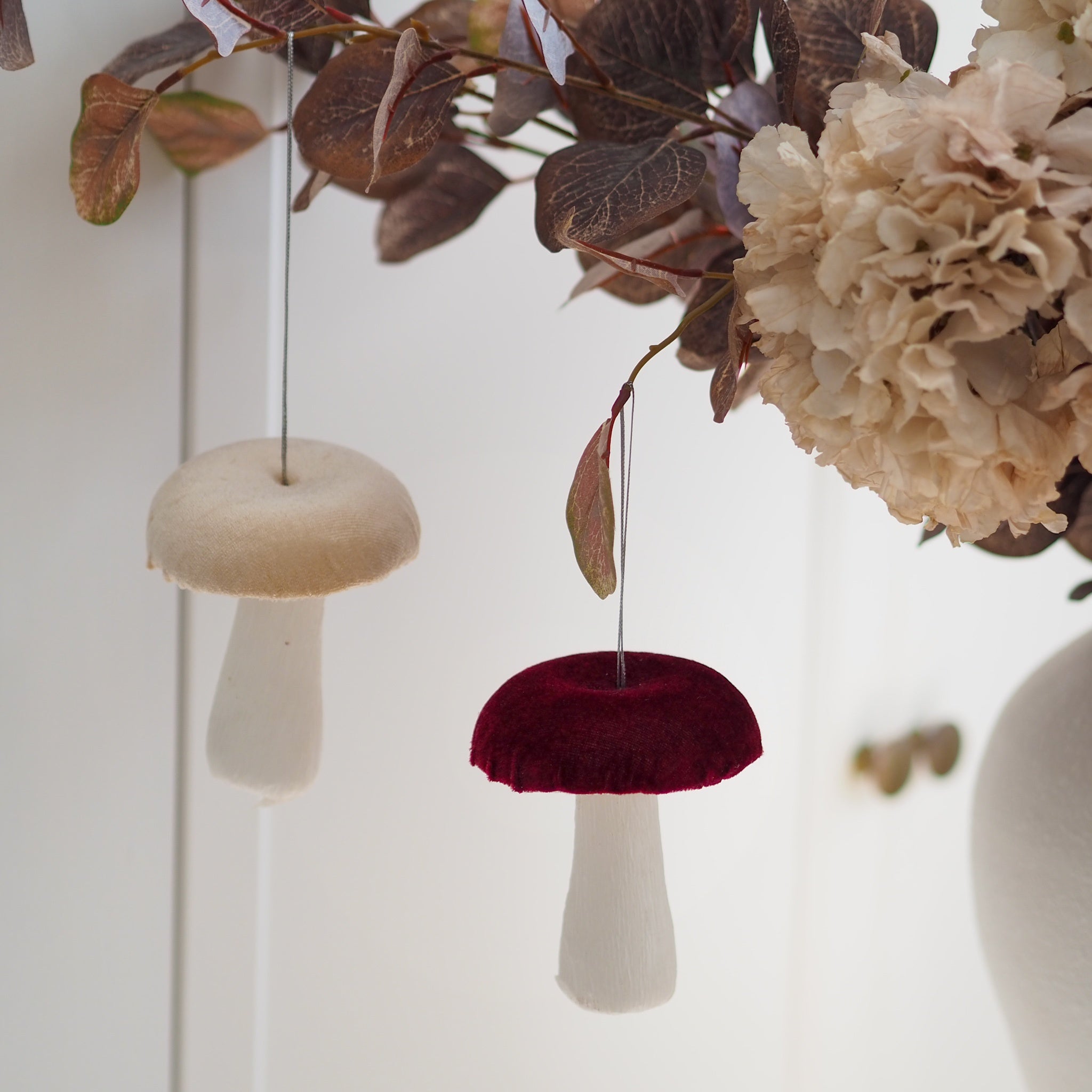 Two mushroom-shaped hanging ornaments, one in creaml and one in burgundy velvet with white stems, suspended in front of a white paneled wall with dried leaves and cream flowers.