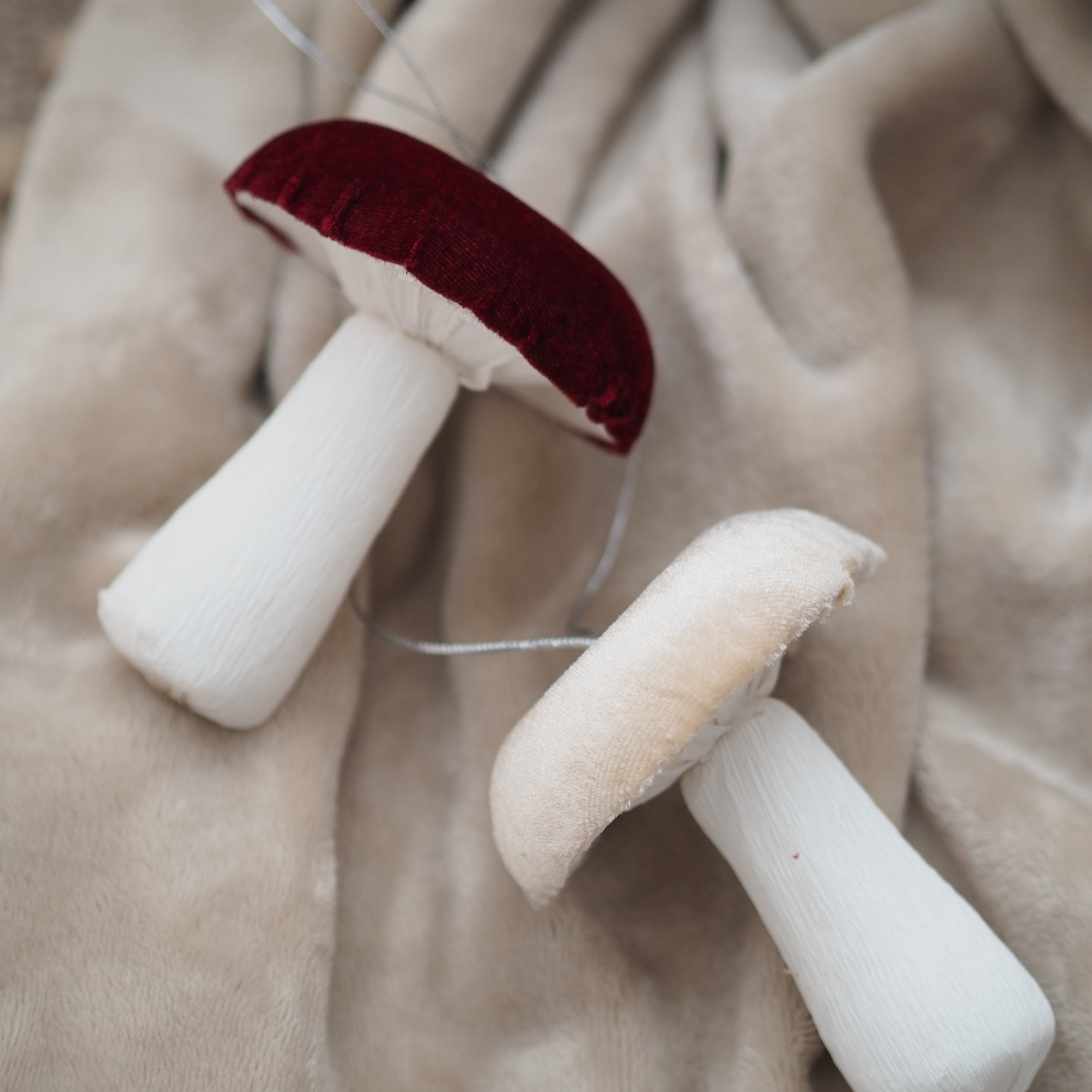 Two plush mushroom decorations with white stems and caps, one with a dark red velvet cap and one with a cream velvet cap, resting on a soft beige fabric background.