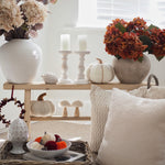 A styled autumn home decor scene featuring a wooden two-tier shelf with white ceramic vases, dried flowers, candles, knitted pumpkins, and decorative pillows in a bright, neutral-toned room.
