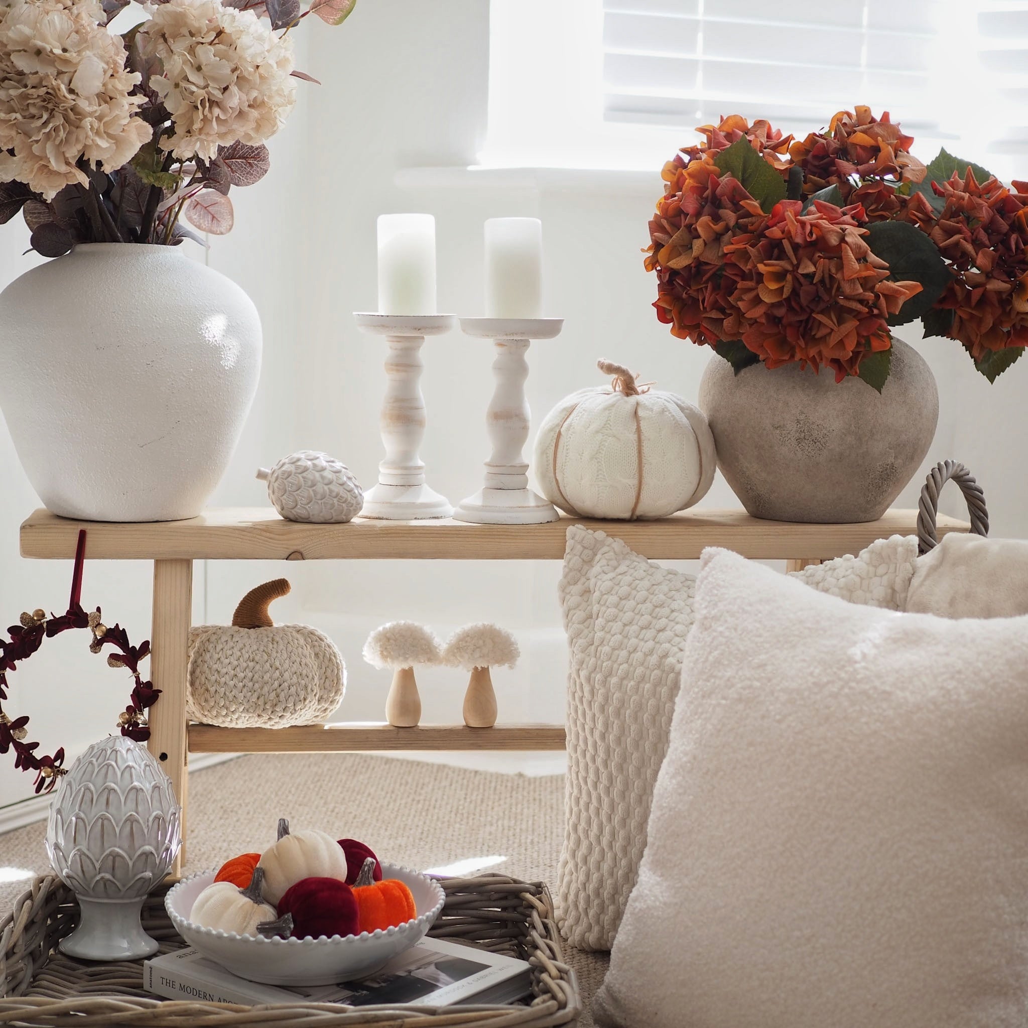 A styled autumn home decor scene featuring a wooden two-tier shelf with white ceramic vases, dried flowers, candles, knitted pumpkins, and decorative pillows in a bright, neutral-toned room.