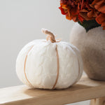 A knitted white faux pumpkin decoration with a textured cable-knit pattern and a natural tan stem, displayed on a wooden shelf next to a vase of orange flowers.