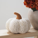 A chunky knitted decorative pumpkin in off-white sits on a light wood surface, with a tan knitted stem and a blurred vase of orange flowers in the background.