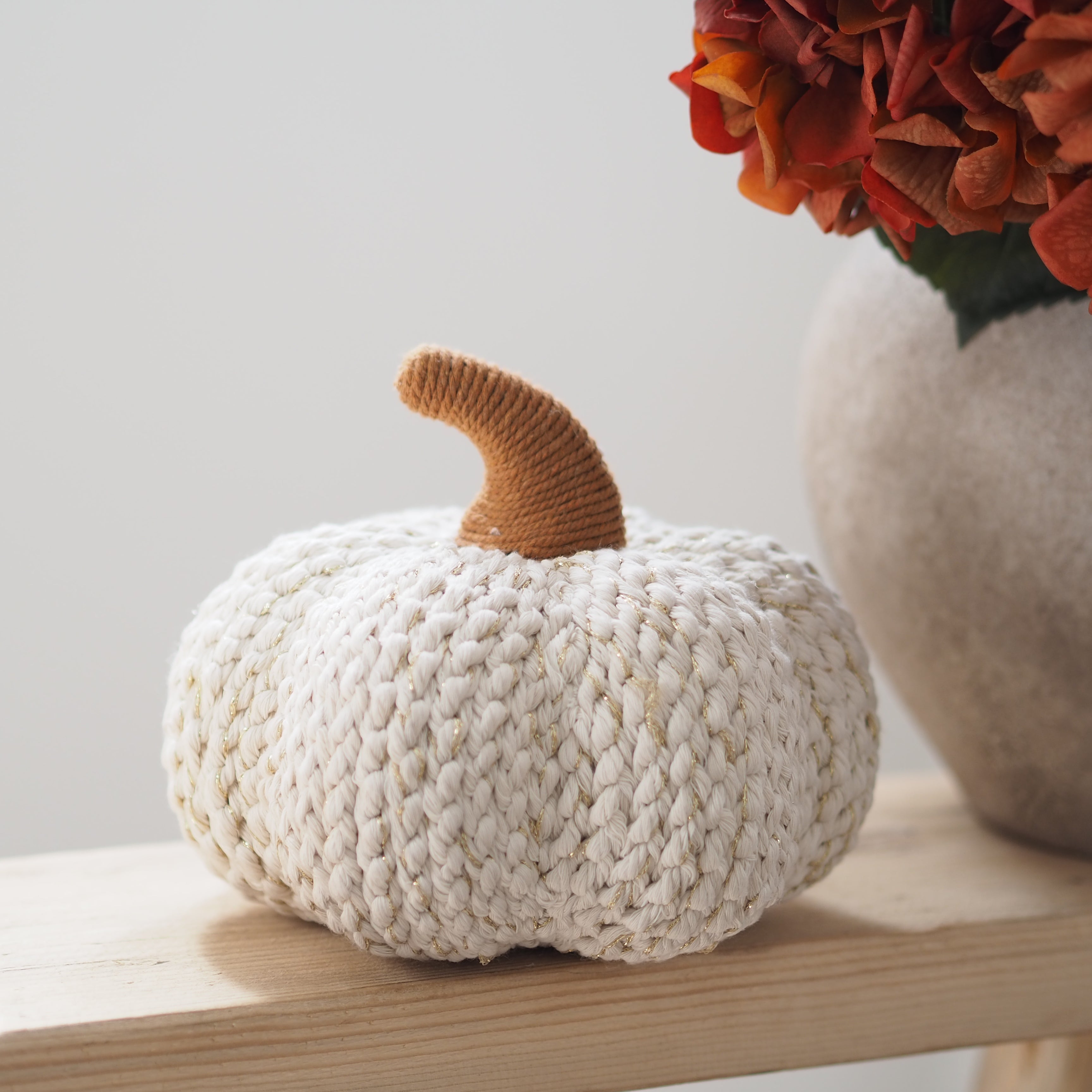 A chunky knitted decorative pumpkin in off-white sits on a light wood surface, with a tan knitted stem and a blurred vase of orange flowers in the background.