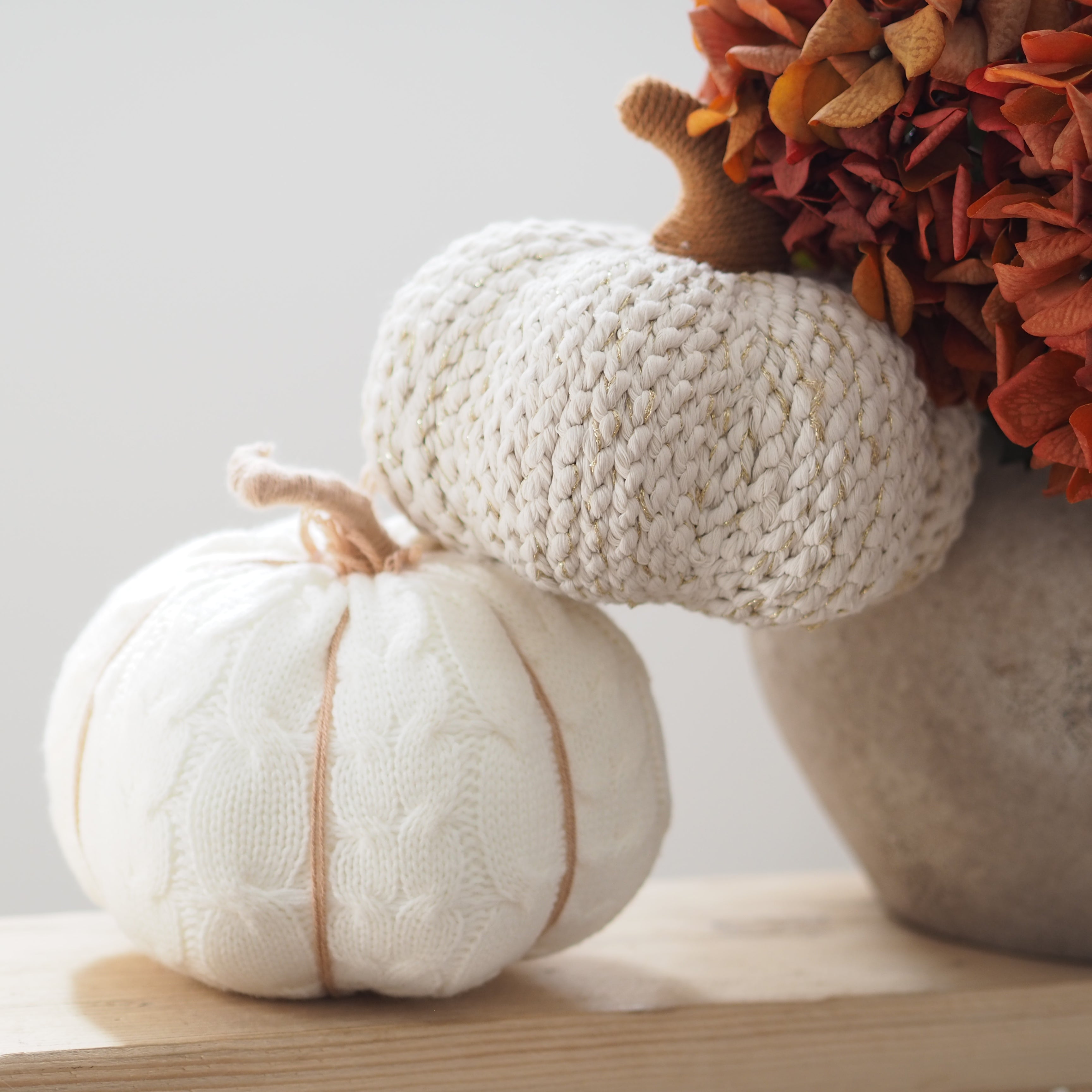 Two knitted decorative pumpkins, one cream and one off-white, styled on a wooden surface next to a vase of dried orange flowers.