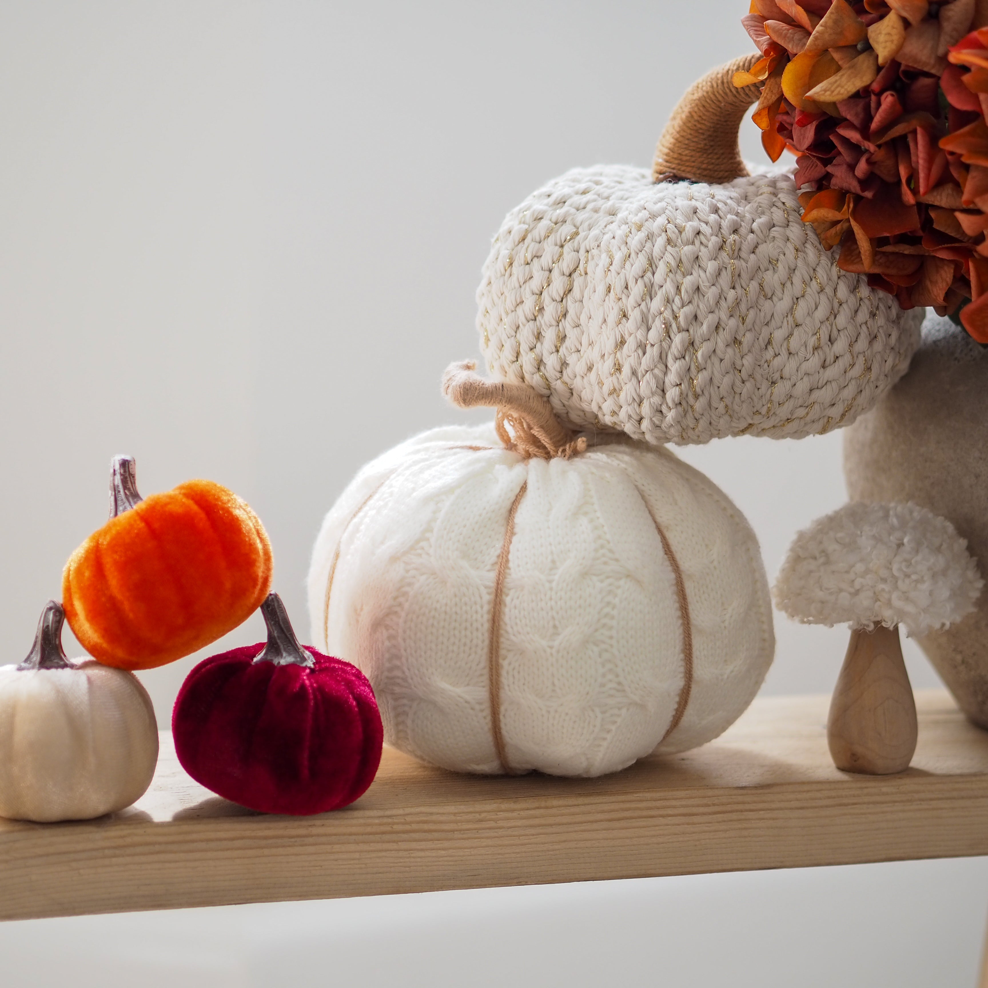 A styled autumn display of decorative pumpkins on a light wood shelf against a neutral off-white background. The collection includes a large cream cable-knit pumpkin, a smaller cream knit pumpkin, a bright orange velvet pumpkin, a deep red velvet pumpkin, a small cream knit pumpkin, a small mushroom-shaped decor piece, and dried orange flowers.