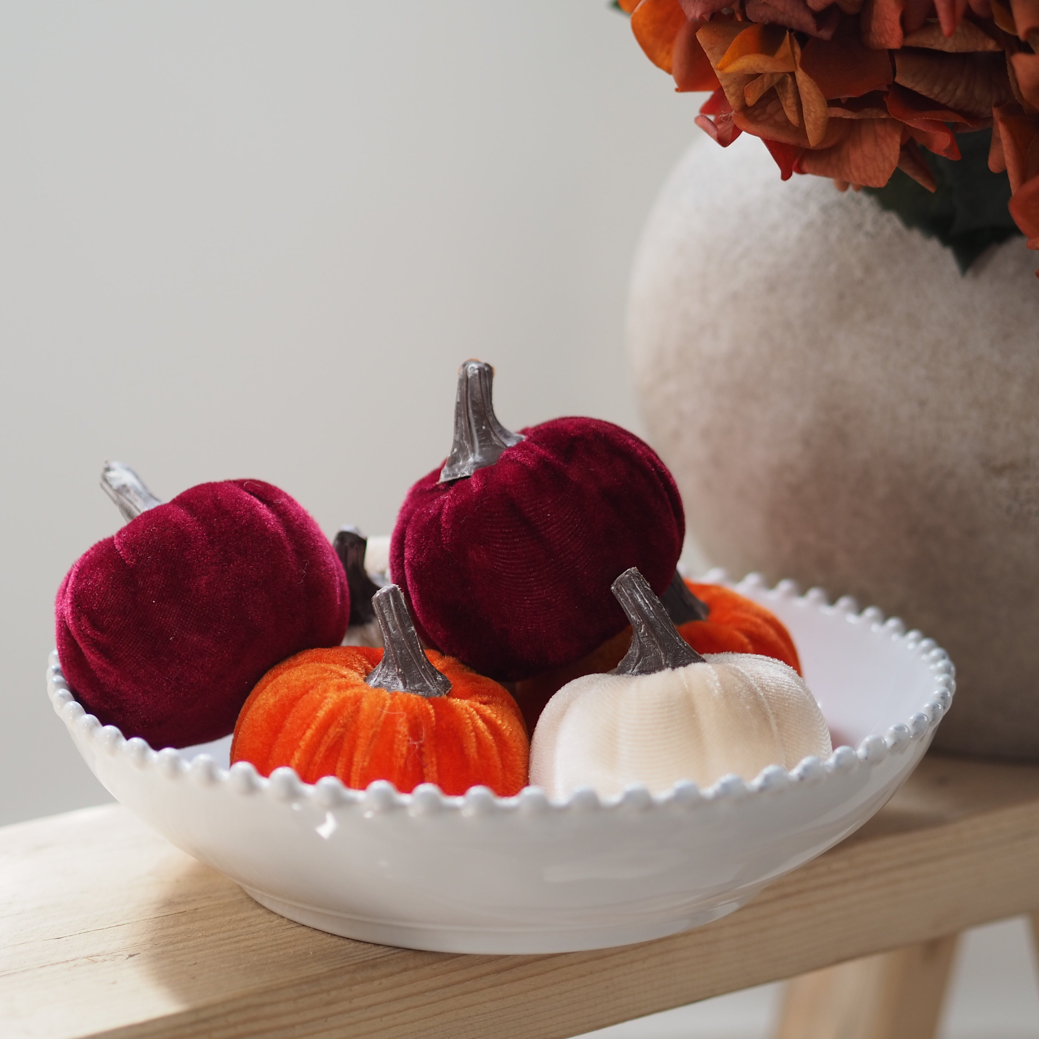 A white ceramic bowl with a scalloped rim holding several decorative velvet pumpkins in burgundy, orange, cream, and dark red, styled on a light wood surface with a blurred floral arrangement in the background.