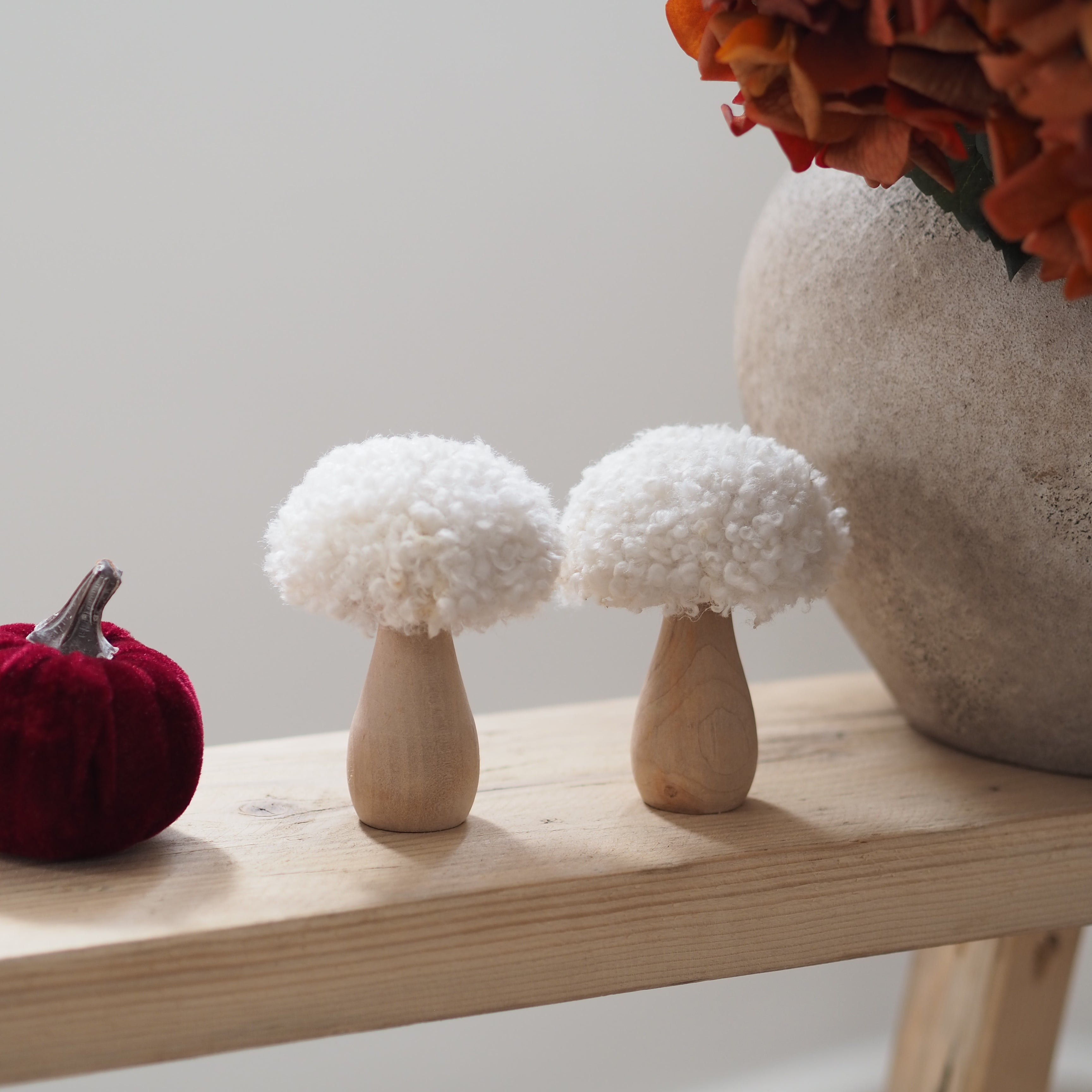 Two decorative toadstool decorations with fluffy white tops and light wood bases, displayed on a light wood shelf beside a burgundy velvet pumpkin and a large textured vase with orange flowers.