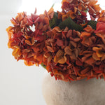 A close-up of a bouquet of artificial hydrangea-style flowers in warm autumn shades of orange, rust, and peach, with a few green leaves and a soft off-white vase against a bright white background.