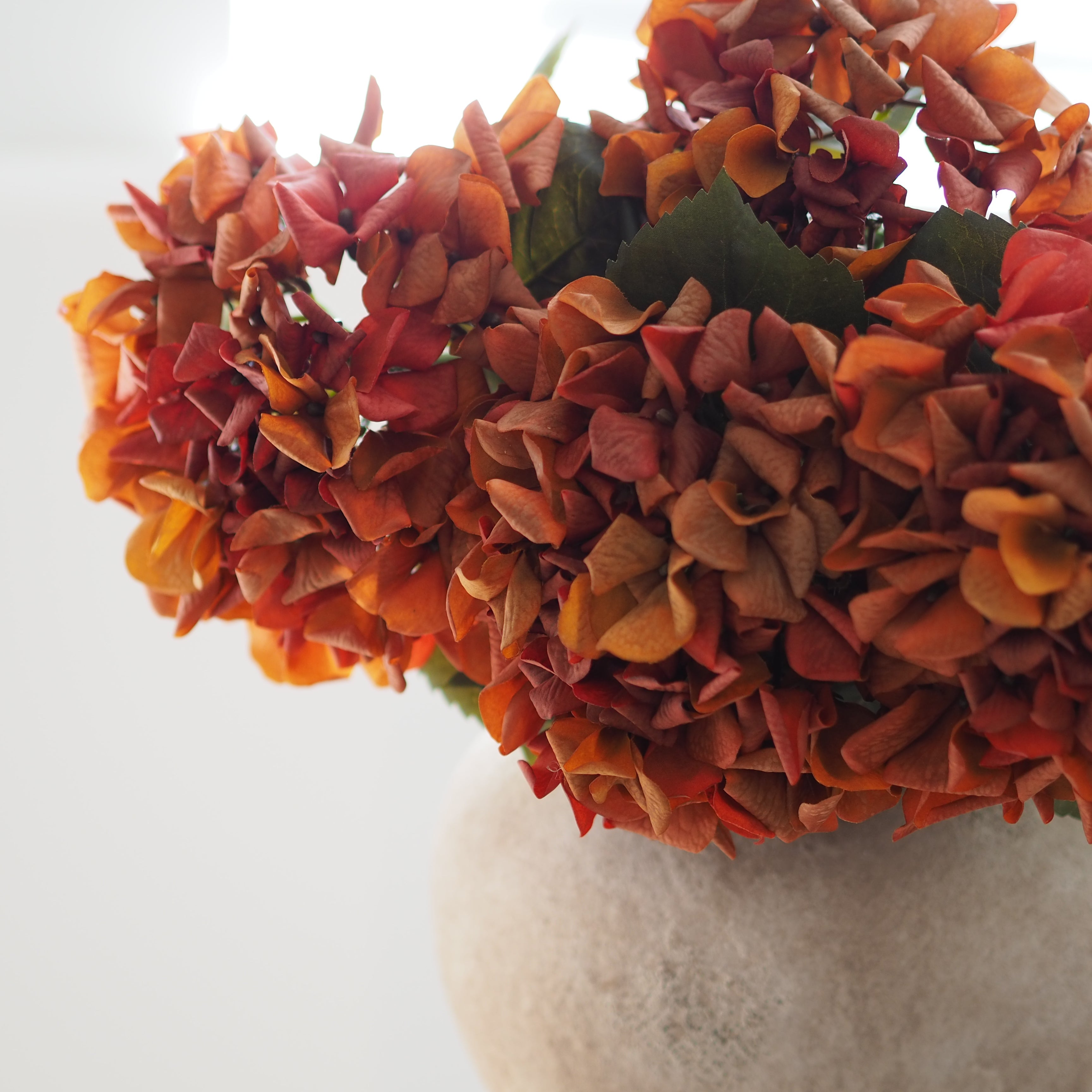 A close-up of a bouquet of artificial hydrangea-style flowers in warm autumn shades of orange, rust, and peach, with a few green leaves and a soft off-white vase against a bright white background.