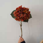 An artificial hydrangea stem with rust-orange petals, green leaves, and a brown branch, held by a hand against a plain light gray wall.