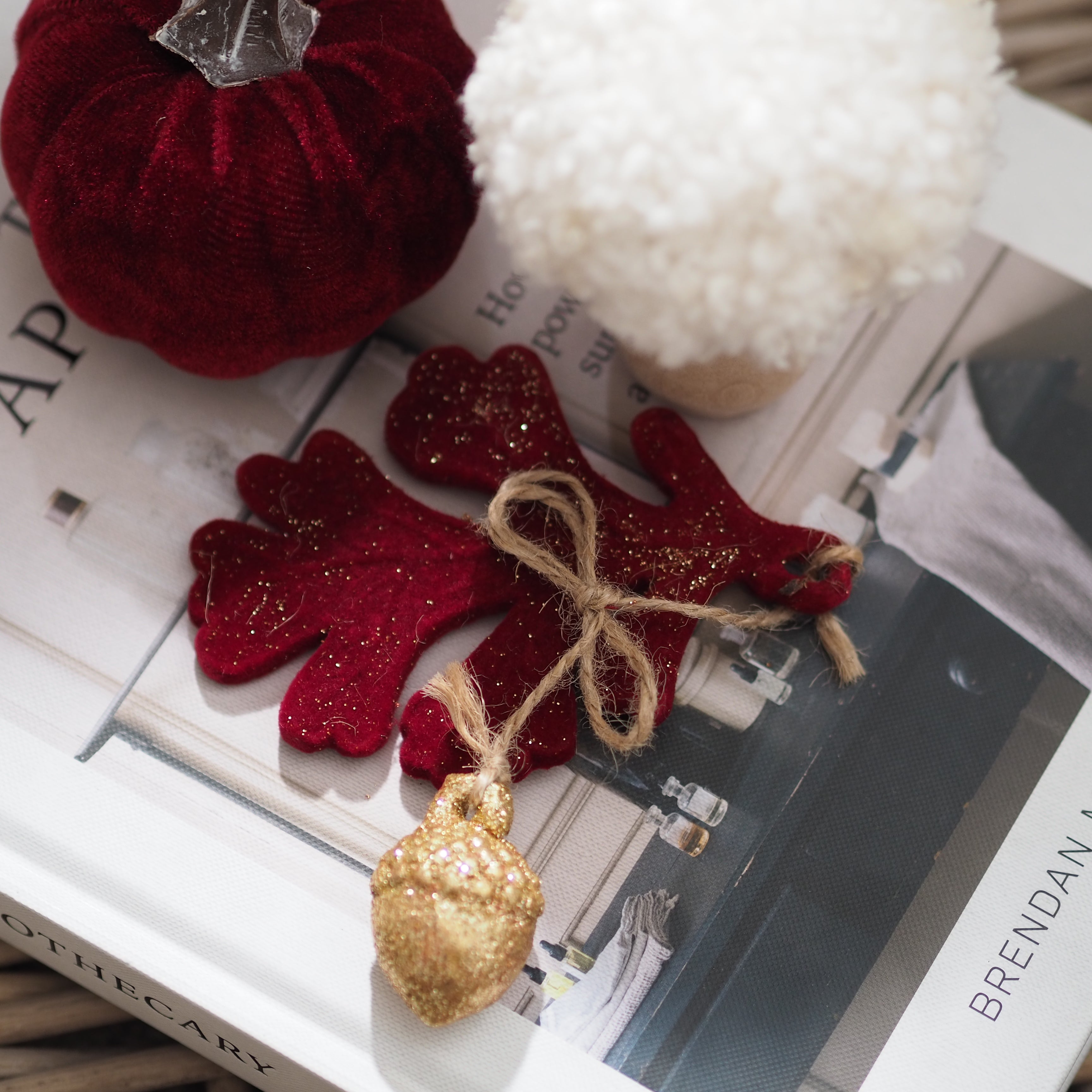 A styled flat-lay of Autumn ornaments on a book, featuring a red velvet pumpkin, a white faux-fur ornament, and burgundy velvet leaf hanging  with gold glitter accents tied with twine and topped with a gold glitter acorn.