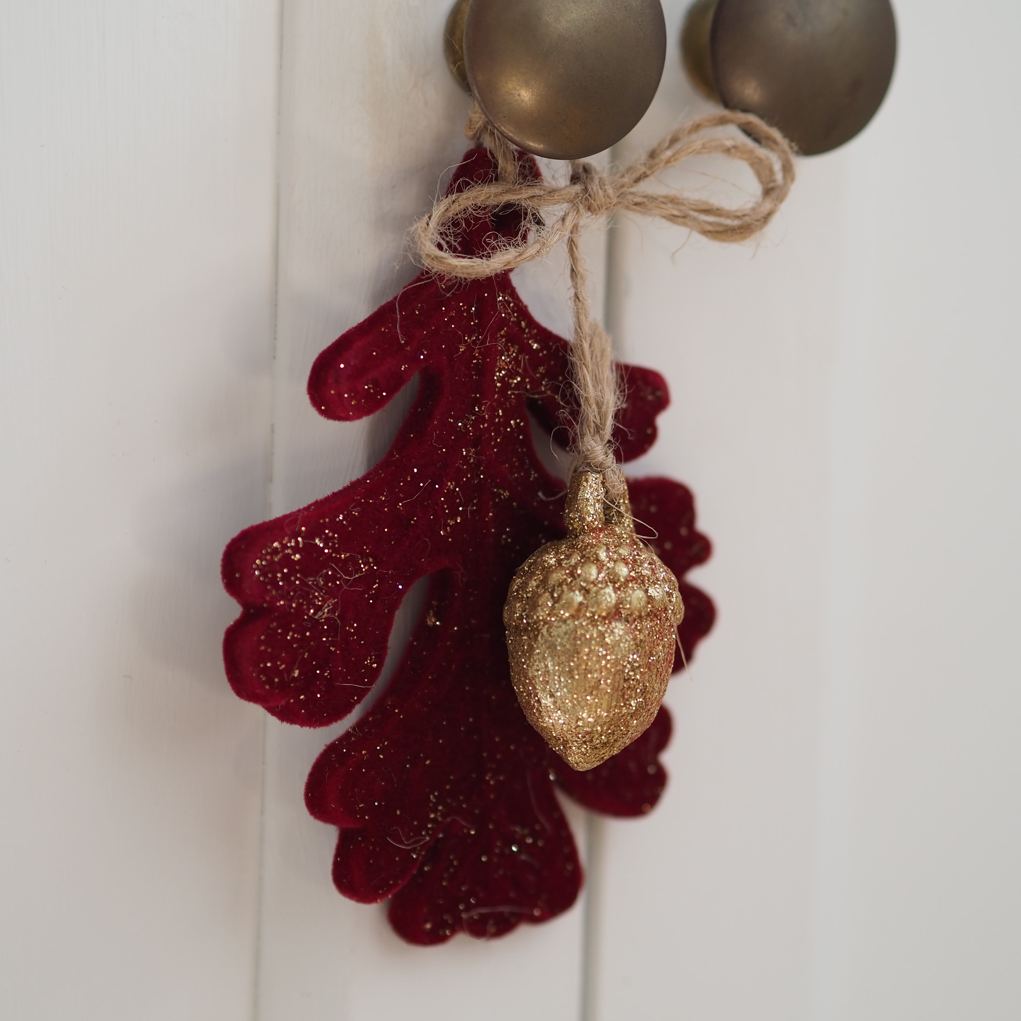 A decorative hanging ornament featuring a glittery deep red leaf and a textured gold acorn charm tied together with natural twine, shown against a white paneled background.