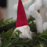A small Christmas gnome decoration with a tall red velvet hat, white beard, and pale pink nose, nestled among green pine branches.