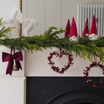 A festive Christmas mantel decorated with a green pine garland, red velvet bow, red heart-shaped wreath, red gnomes, and a white fluffy ornament against a white fireplace.