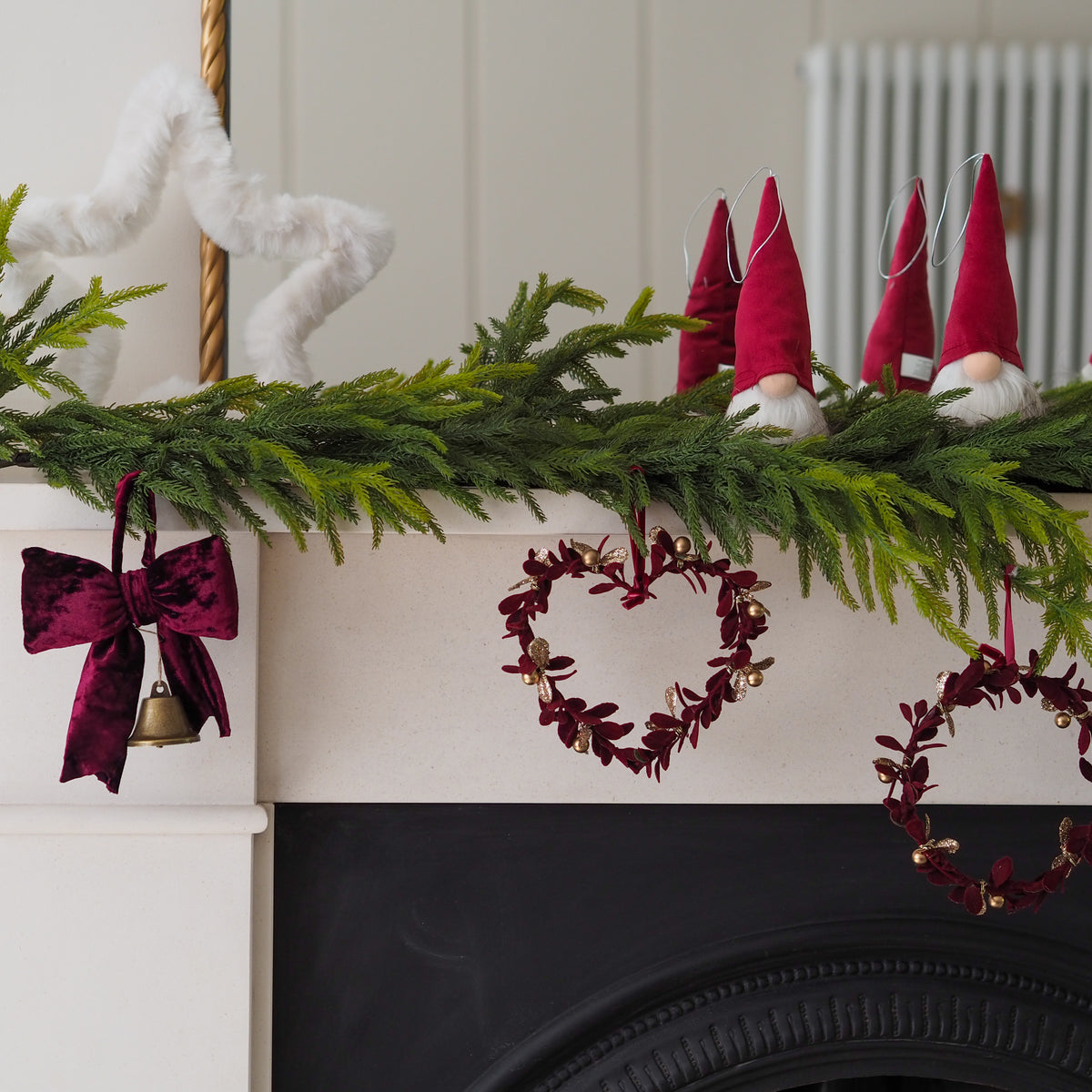 A festive Christmas mantel decorated with a green pine garland, red velvet bow, red heart-shaped wreath, red gnomes, and a white fluffy ornament against a white fireplace.