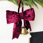 A deep burgundy velvet Christmas ornament shaped like a bow with a small gold bell hanging from the center, suspended from a green pine branch against a white background.