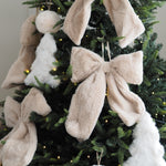 Fuzzy plush Christmas ornaments, including large beige bunny ears and bows, white fluffy angel figures, and a white bunny tail, hanging on a dark green Christmas tree with warm string lights.
