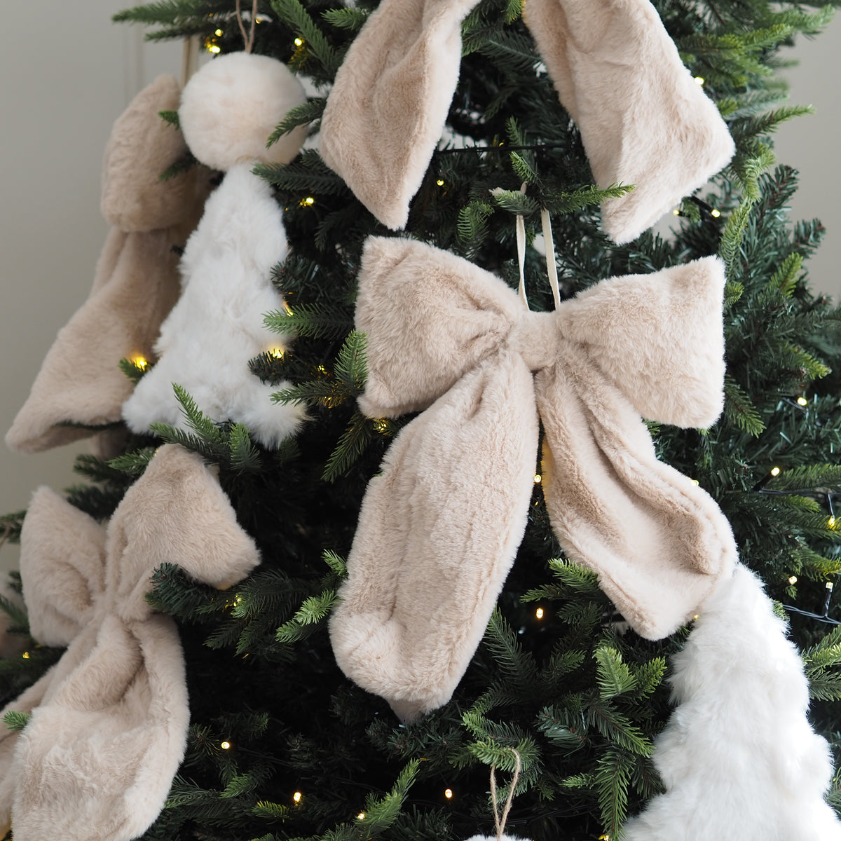 Fuzzy plush Christmas ornaments, including large beige bunny ears and bows, white fluffy angel figures, and a white bunny tail, hanging on a dark green Christmas tree with warm string lights.