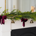 A green pine garland draped over a white fireplace mantel, decorated with two deep burgundy velvet bows and small hanging brass bells.