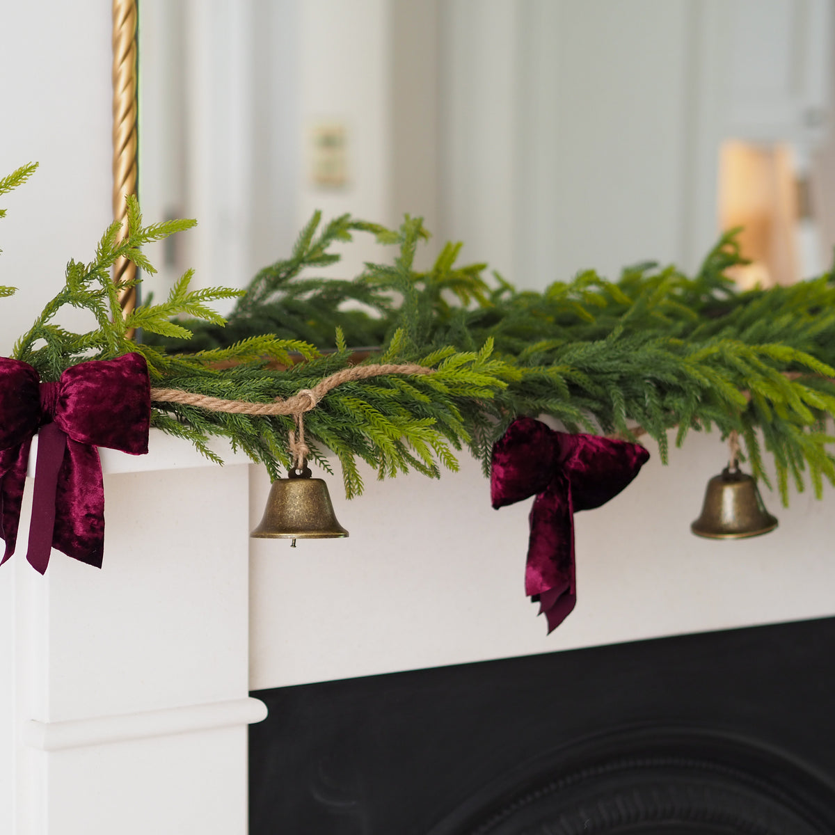 A green pine garland draped over a white fireplace mantel, decorated with two deep burgundy velvet bows and small hanging brass bells.