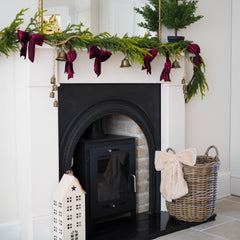 A styled Christmas fireplace scene with a green pine garland draped across a white mantel, accented with dark burgundy velvet bows, small gold bells, and a potted green tree. A black wood-burning stove sits in the fireplace, with a wicker basket holding a cream throw and a white decorative house ornament on the hearth.