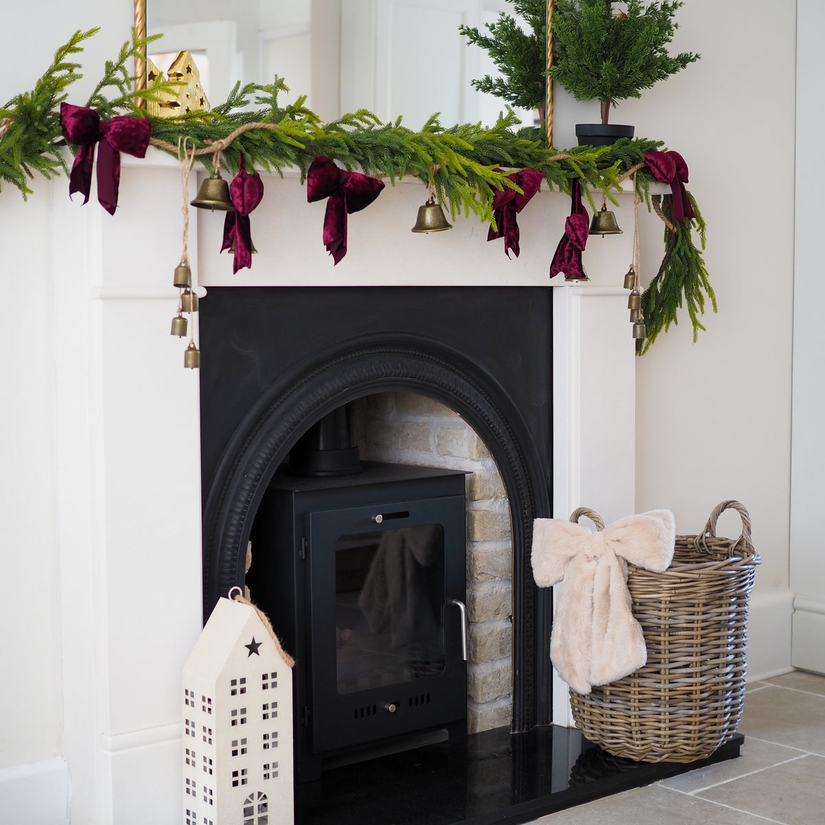 A styled Christmas fireplace scene with a green pine garland draped across a white mantel, accented with dark burgundy velvet bows, small gold bells, and a potted green tree. A black wood-burning stove sits in the fireplace, with a wicker basket holding a cream throw and a white decorative house ornament on the hearth.