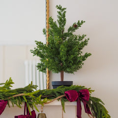 A small artificial Christmas tree in a dark pot, displayed on a mantelpiece above a green garland with burgundy velvet bows.