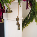 A close-up of a white fireplace mantel decorated with green pine garland, deep burgundy velvet ribbons, and hanging brass bell ornaments.