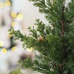 Close-up of green artificial pine or cypress tree branches with small pinecones, shown against a softly blurred warm indoor background with bokeh lights.