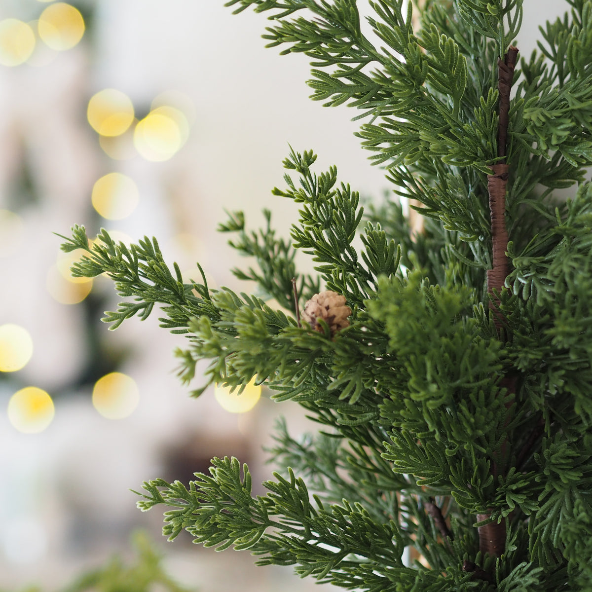 Close-up of green artificial pine or cypress tree branches with small pinecones, shown against a softly blurred warm indoor background with bokeh lights.