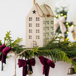 A beige wooden house-shaped hanging ornament with a star on the roof, displayed on a holiday garland with green pine branches, burgundy velvet bows, and small bells.