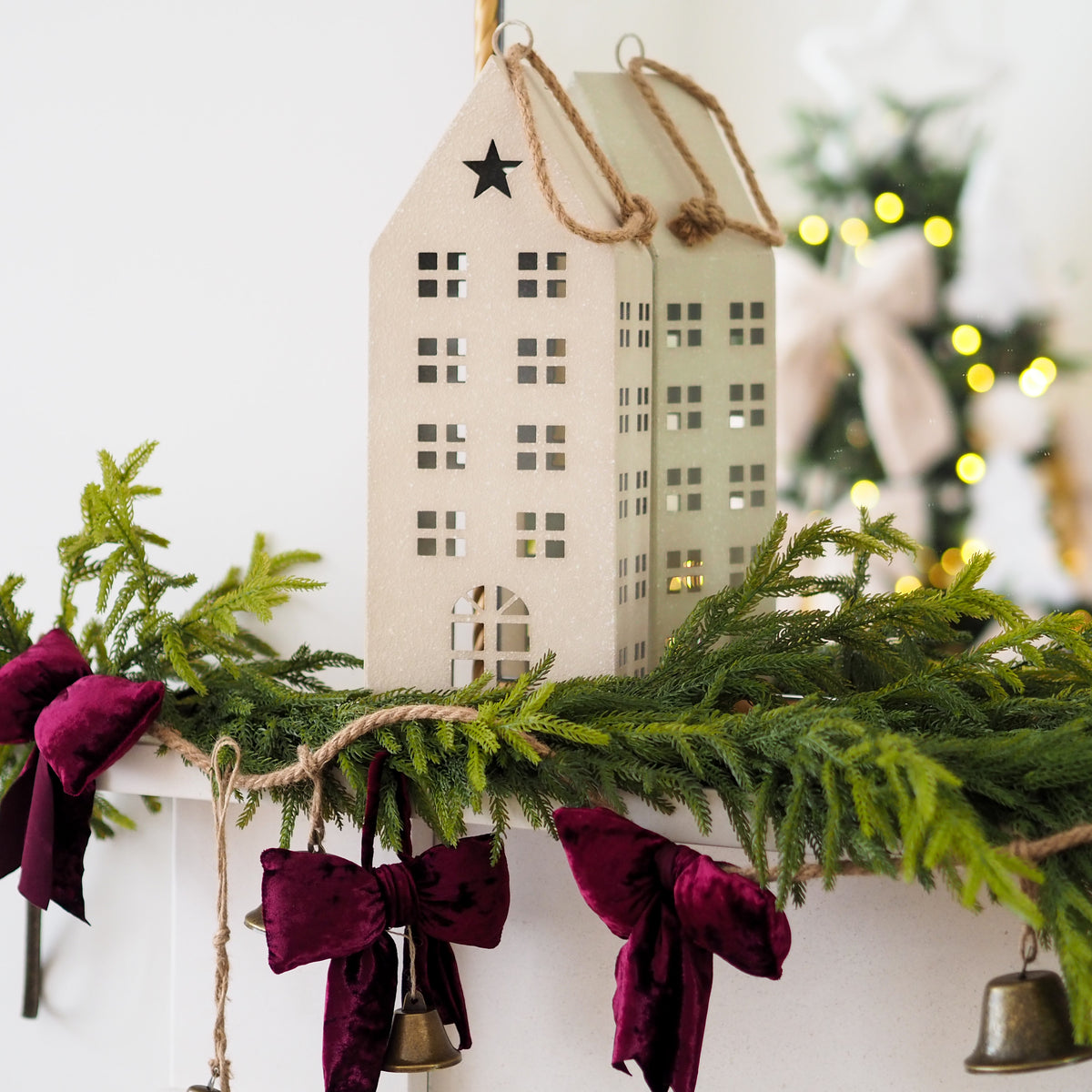 A beige wooden house-shaped hanging ornament with a star on the roof, displayed on a holiday garland with green pine branches, burgundy velvet bows, and small bells.