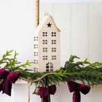 A small beige house-shaped hanging ornament with a star on the roof, displayed on a mantel with green garland, burgundy velvet bows, and small gold bells.