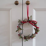A festive Christmas wreath hanging on a white paneled door. The wreath is decorated with green leaves, pinecones, small red berries, and a red velvet ribbon bow with a red hanging loop.