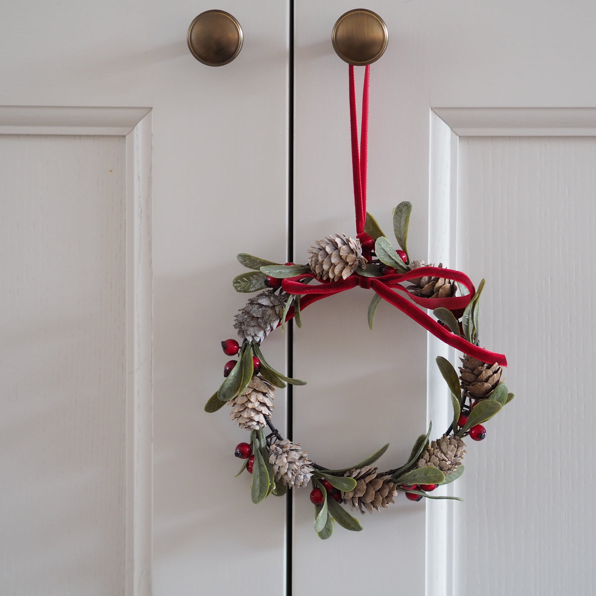 A festive Christmas wreath hanging on a white paneled door. The wreath is decorated with green leaves, pinecones, small red berries, and a red velvet ribbon bow with a red hanging loop.