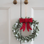 A festive felt Christmas wreath hanging on a white paneled door. The wreath is made of textured sage green leaves with a large, fluffy red bow at the top center and small white pom-pom berries throughout.