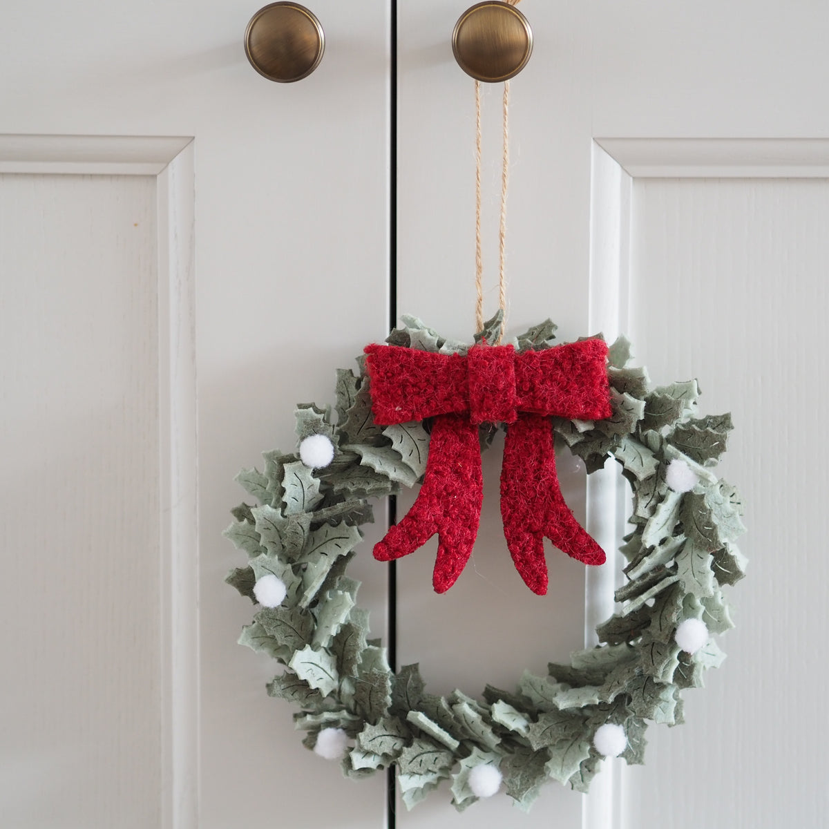 A festive felt Christmas wreath hanging on a white paneled door. The wreath is made of textured sage green leaves with a large, fluffy red bow at the top center and small white pom-pom berries throughout.