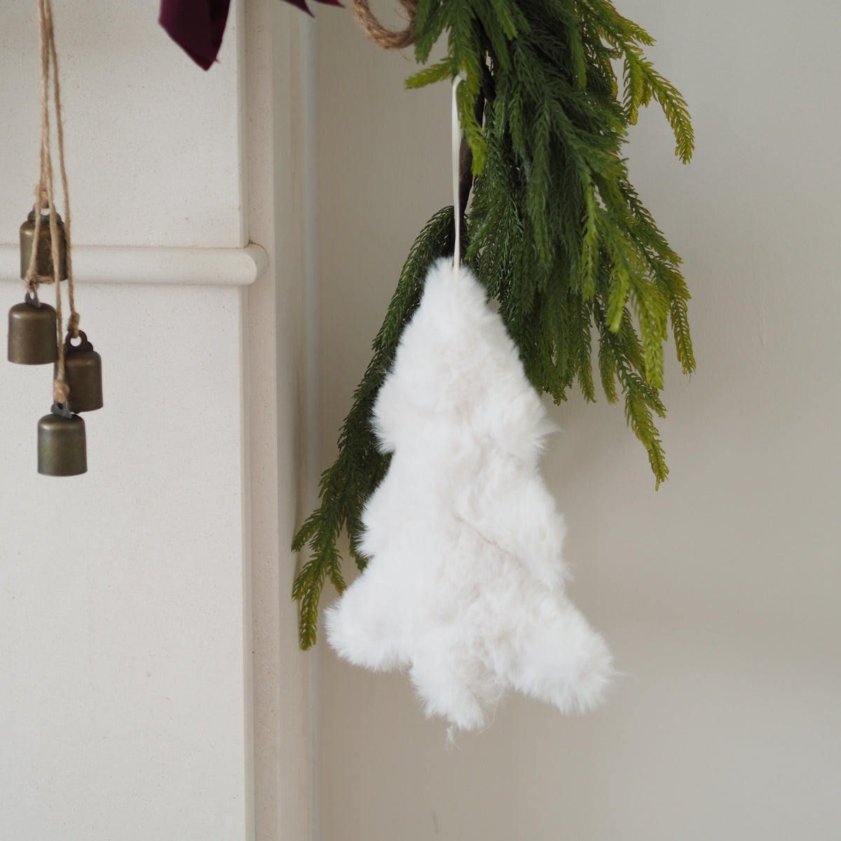 A fluffy white Christmas tree ornament hanging from a garland of green pine branches against a light cream wall.